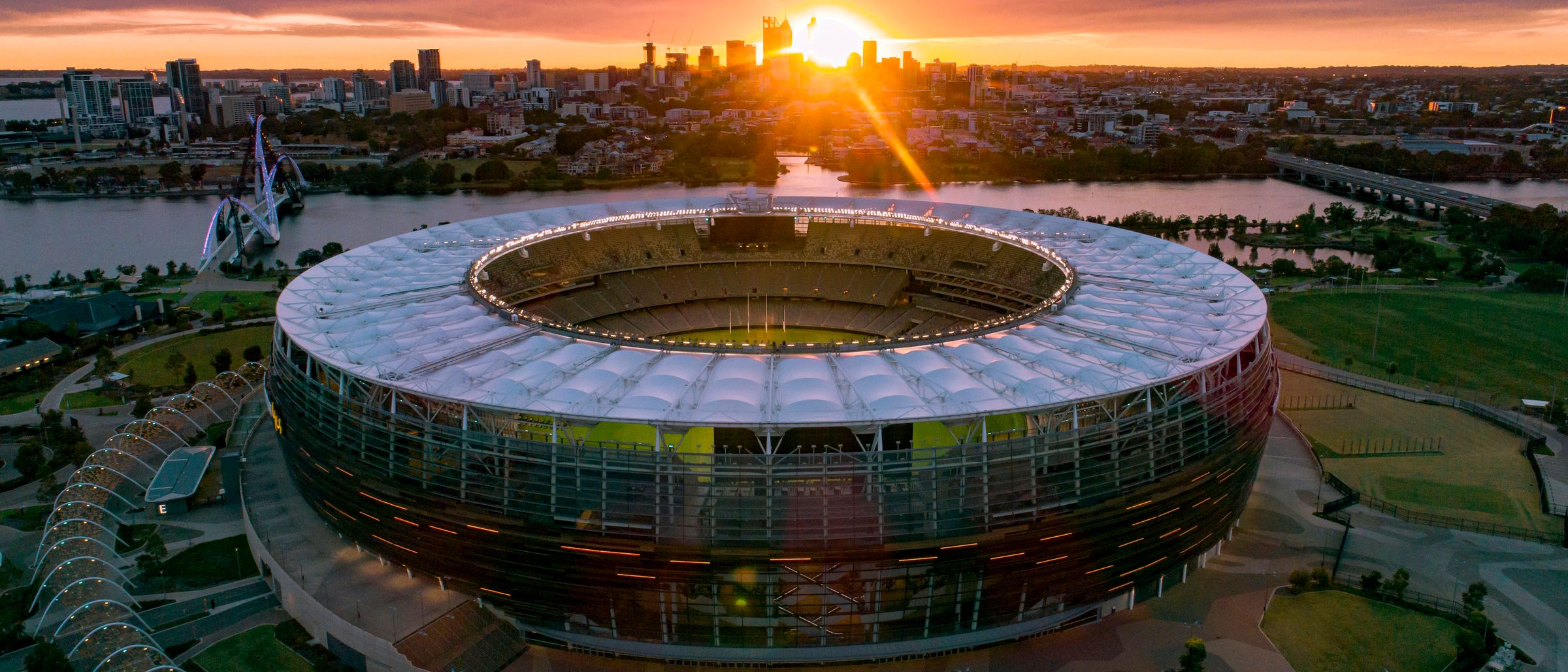 Aerial view of a large oval stadium at dusk beside a river, with a city skyline behind