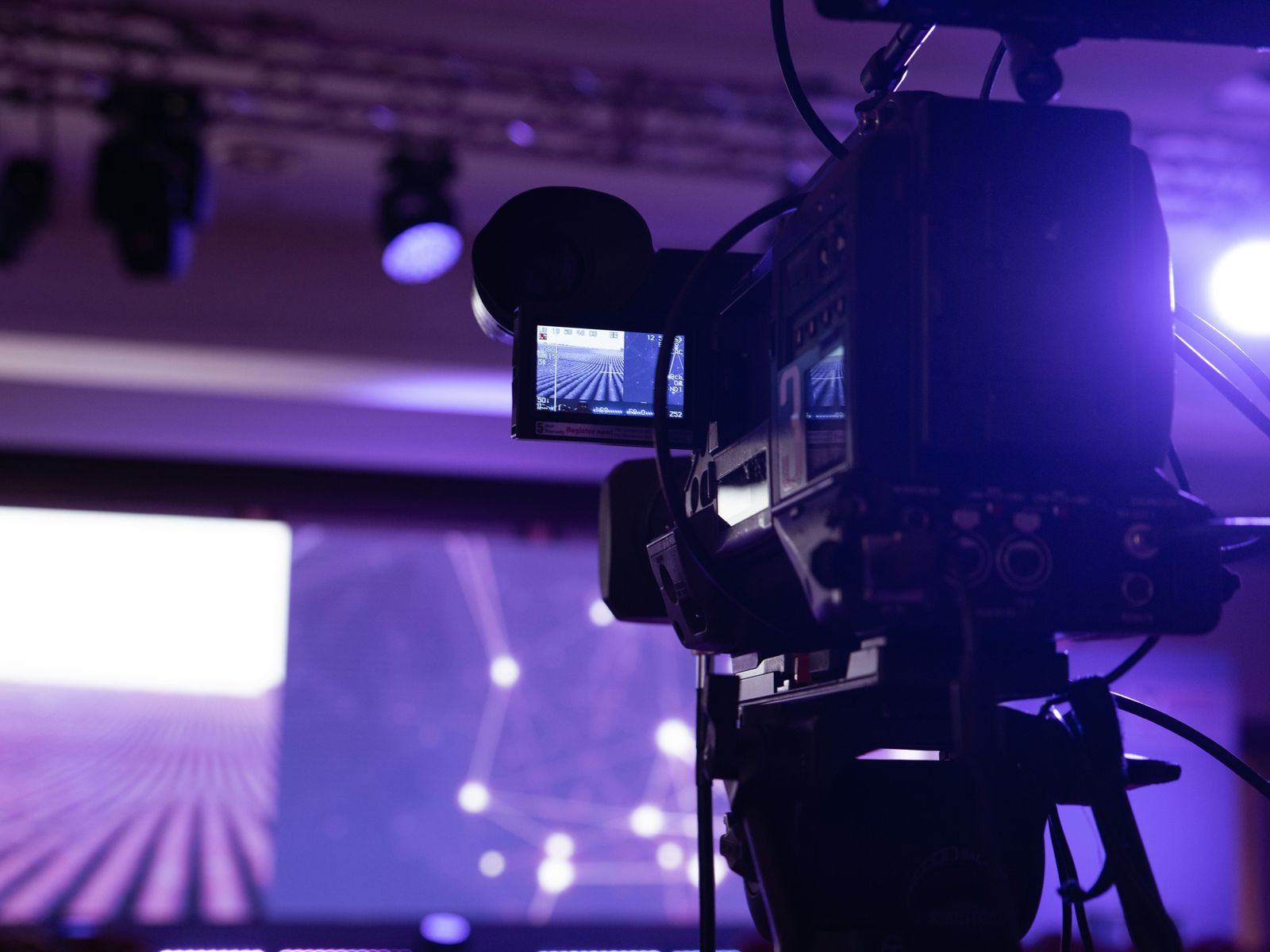 Broadcast video camera on a tripod facing a purple-lit event stage
